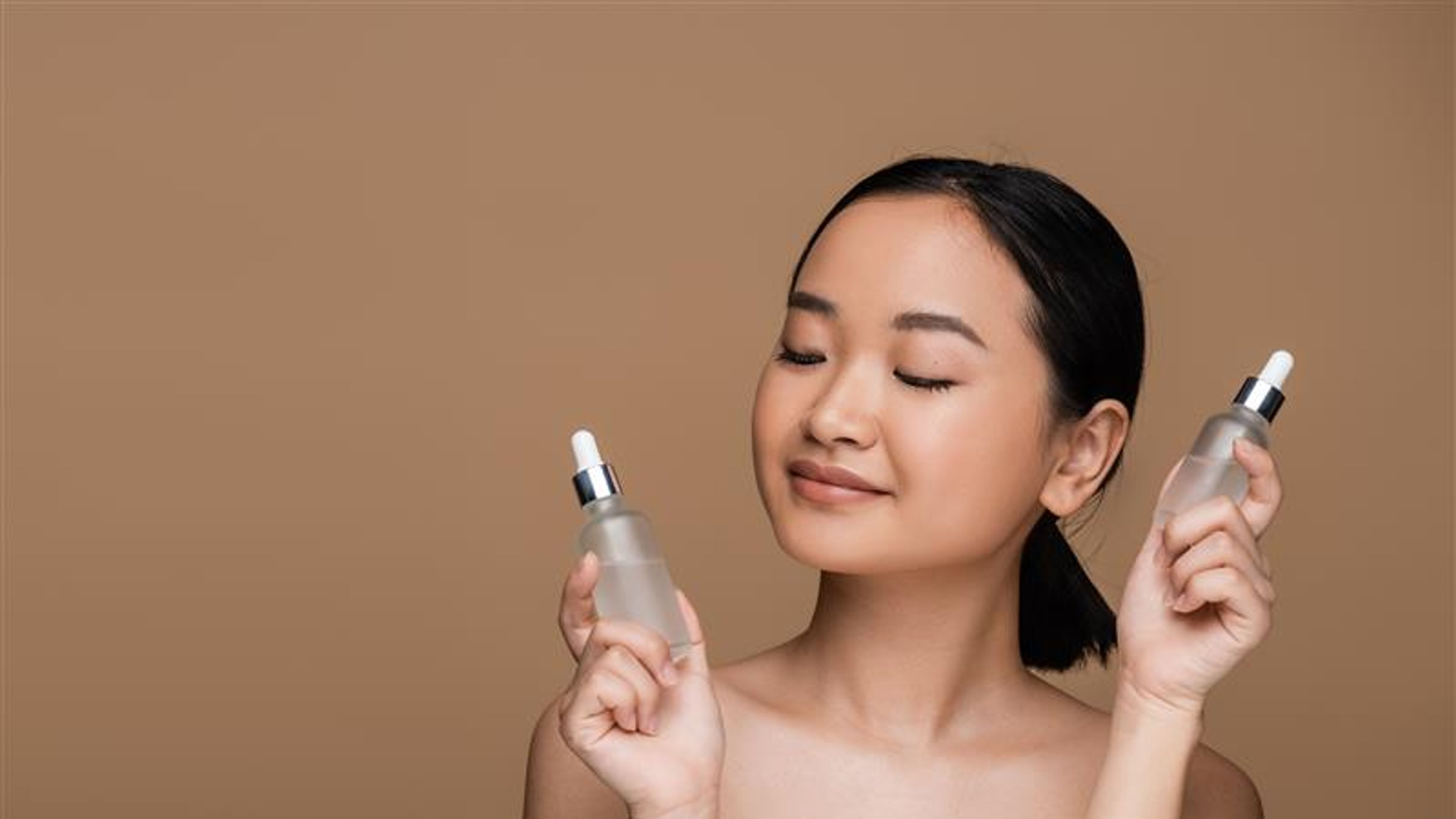 Pretty asian woman with naked shoulders holding bottles of serums isolated on brown